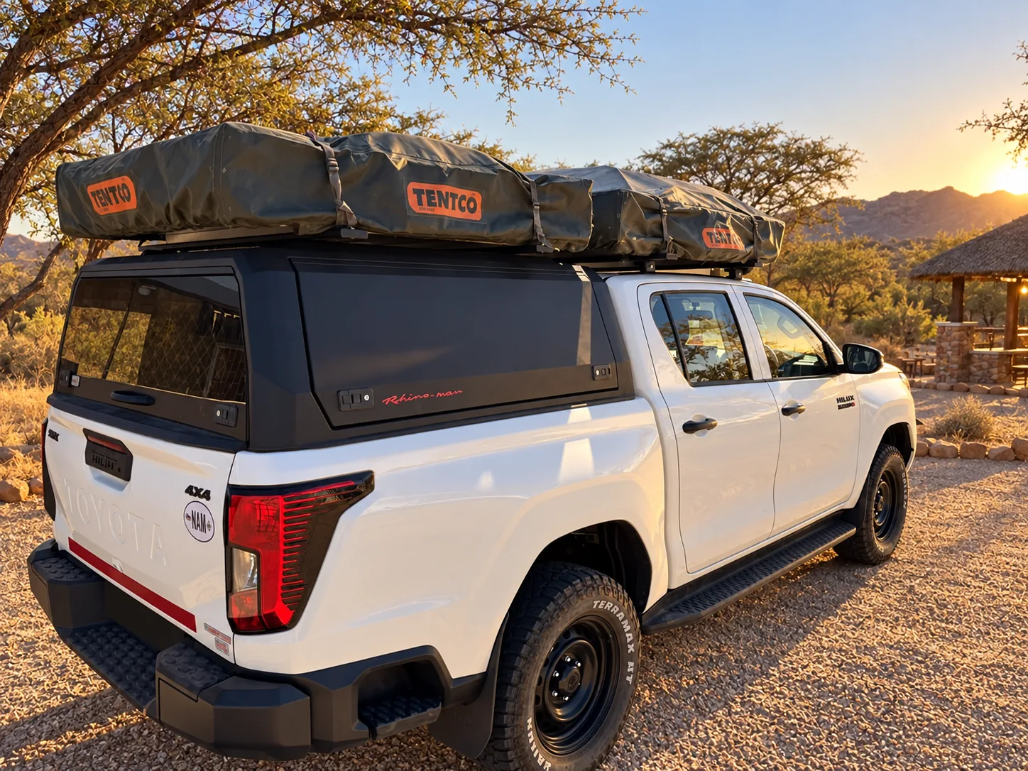 White Toyota Hilux double cab with canopy and two folded rooftop tents, rear three-quarter view, at a lodge