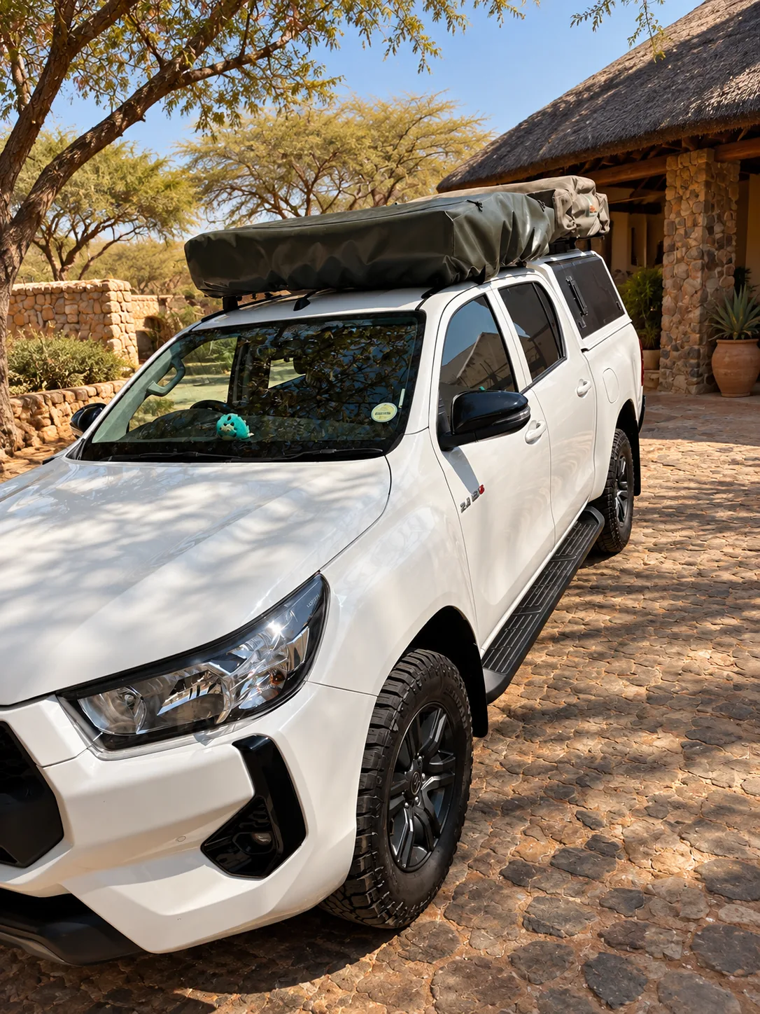 White Toyota Hilux double cab with one folded rooftop tent, front three-quarter view, parked at a thatched lodge