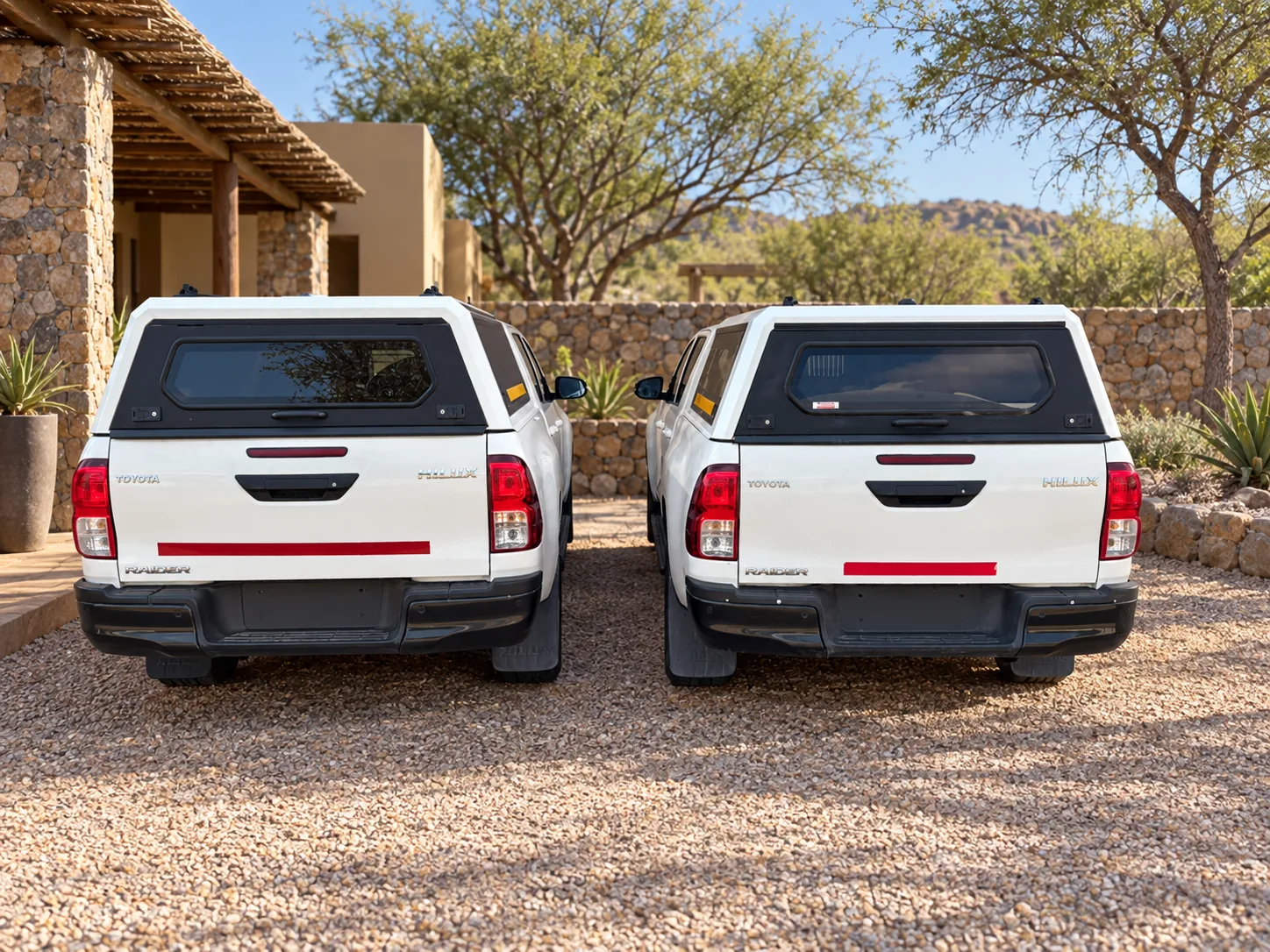 Two white Toyota Hilux double cabs with canopies, rear view, parked at a stone lodge
