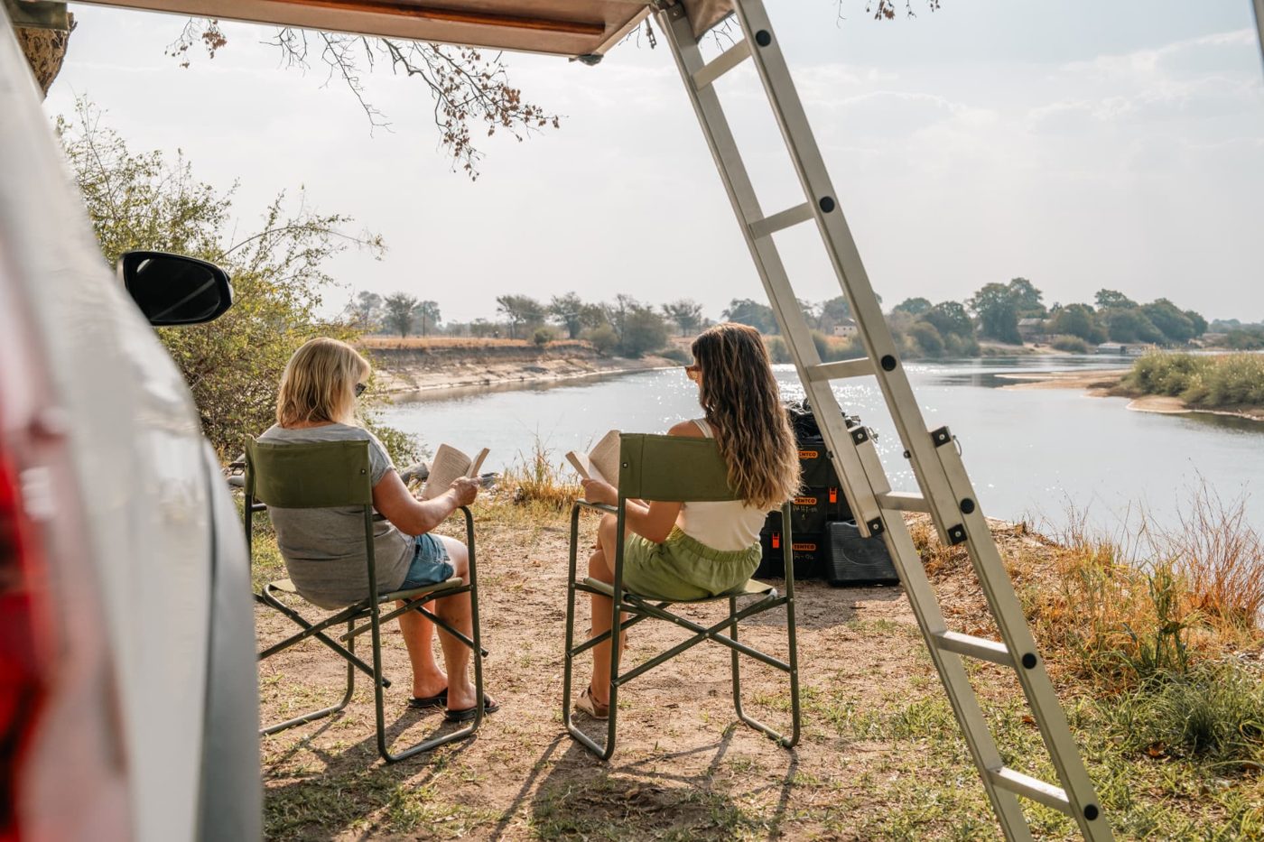 Two travellers reading in camp chairs under the awning beside a river in Namibia