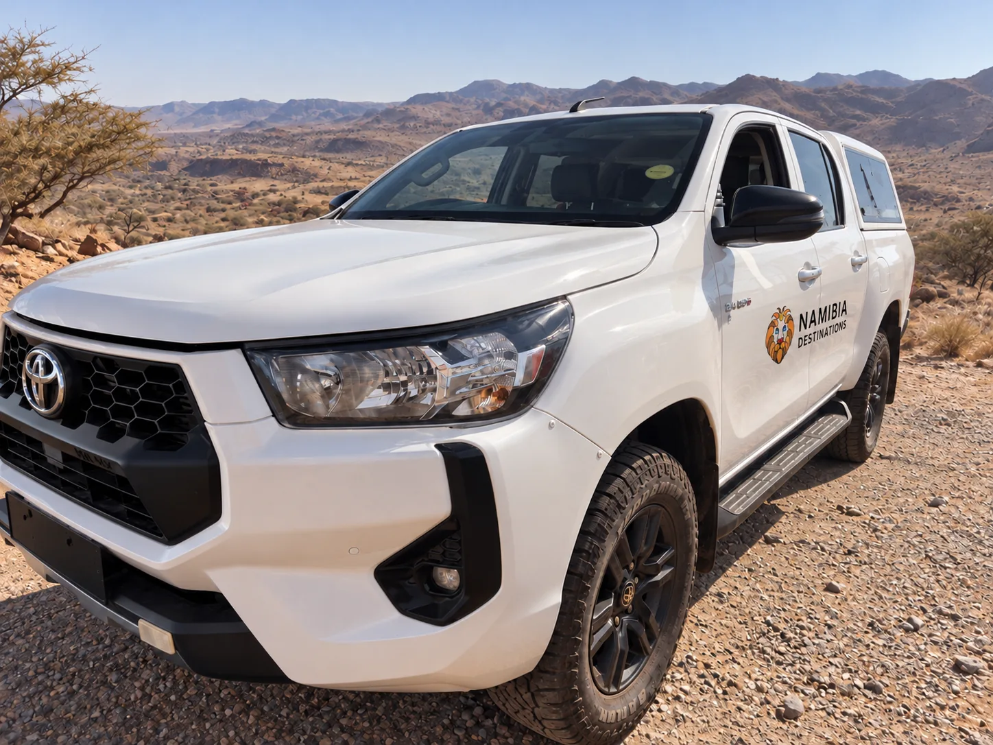White Toyota Hilux 2.4 double cab with canopy, front three-quarter view, in an arid mountain landscape