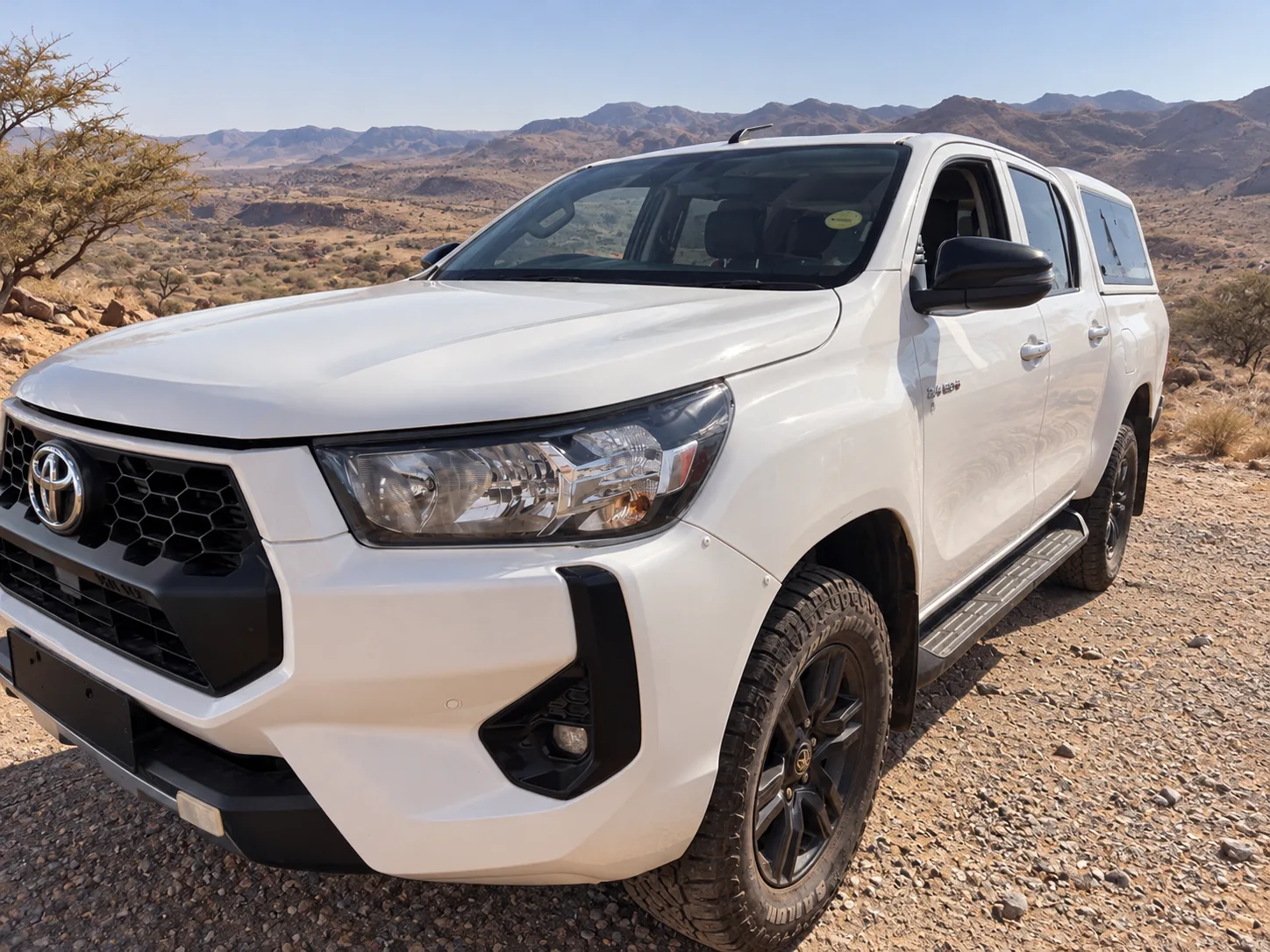 White Toyota Hilux double cab with canopy, front three-quarter view, in an arid mountain landscape